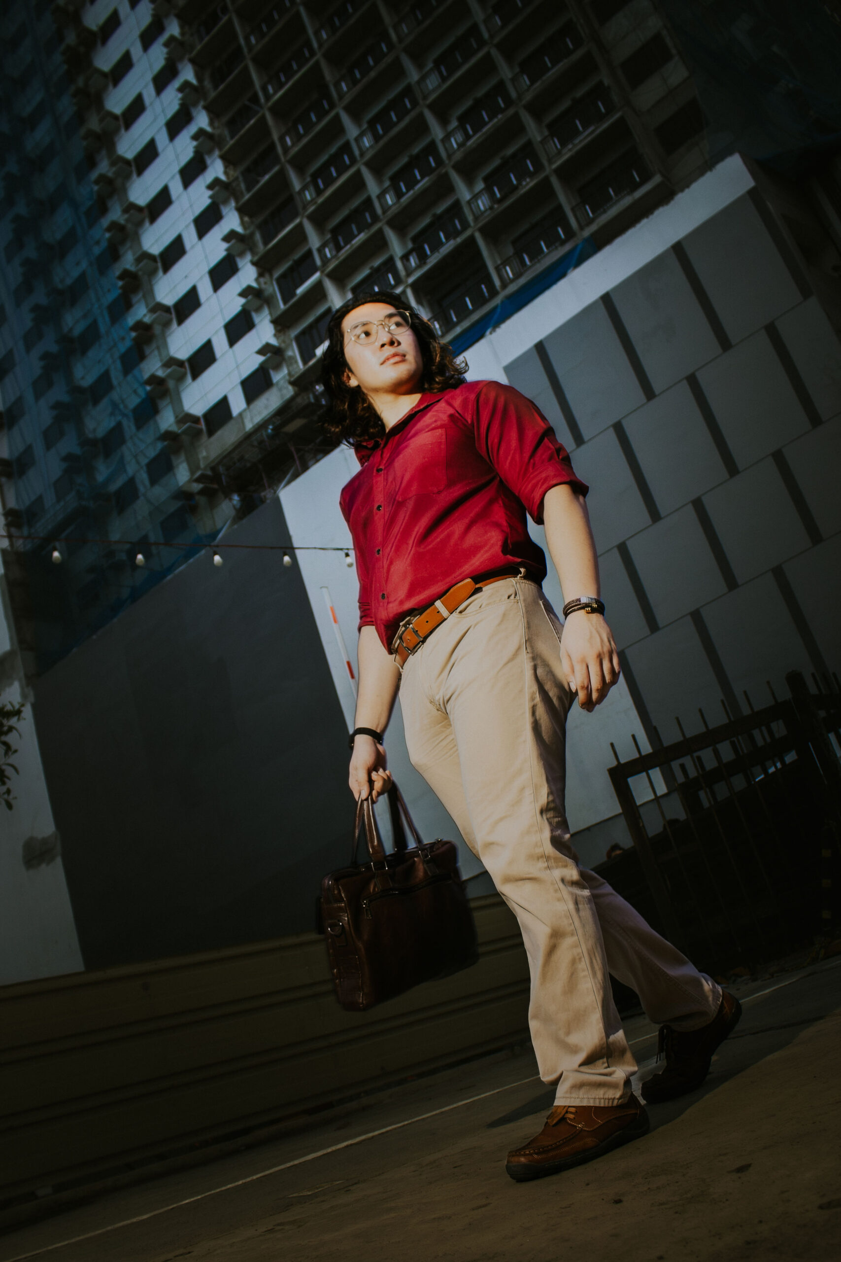 Low-angle shot of Liam walking with a leather bag past a tower block
